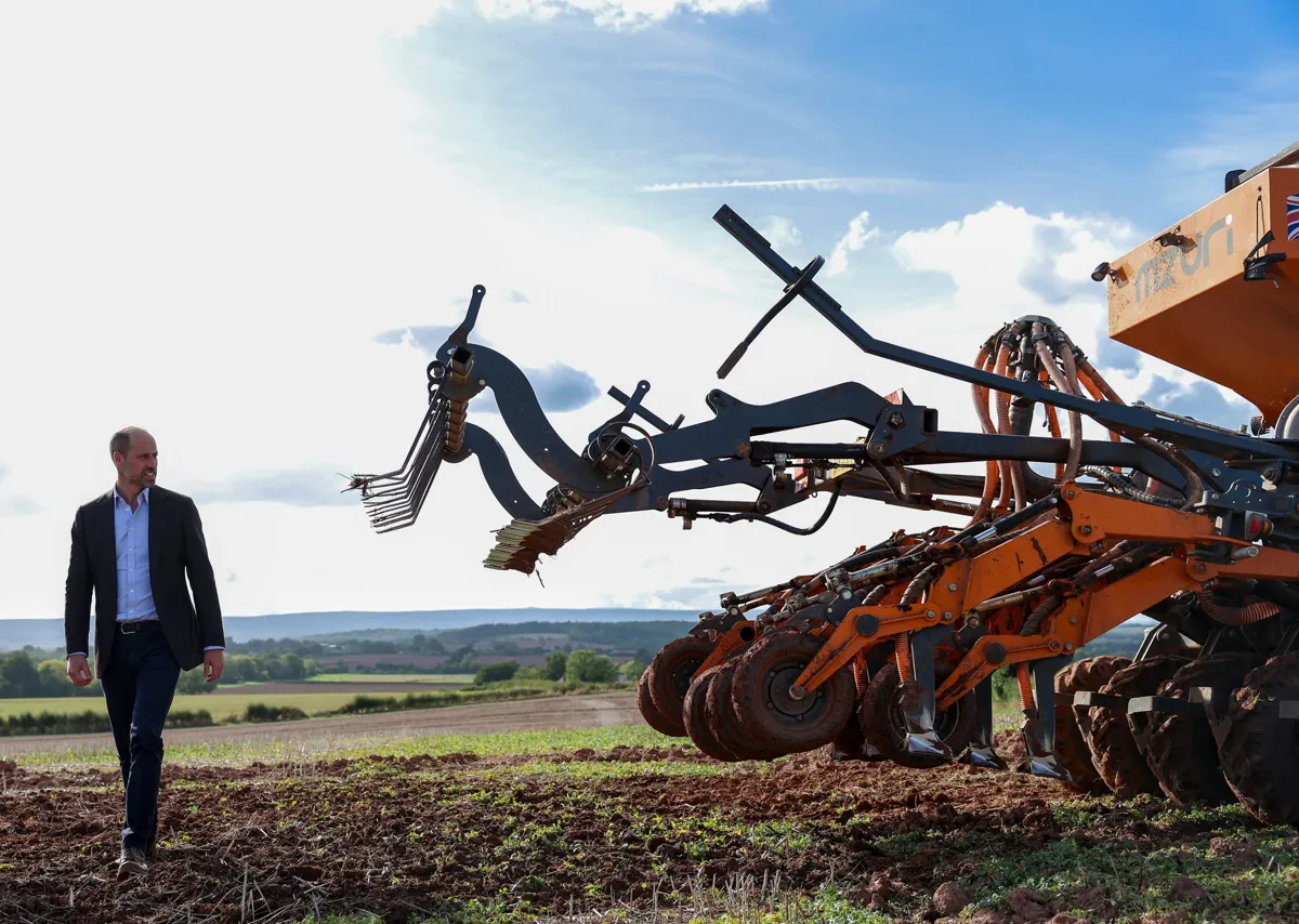 The Duke of Cornwall walks across a drilled field at Monkhall Farm beside an orange seed drill, with the Herefordshire hills behind