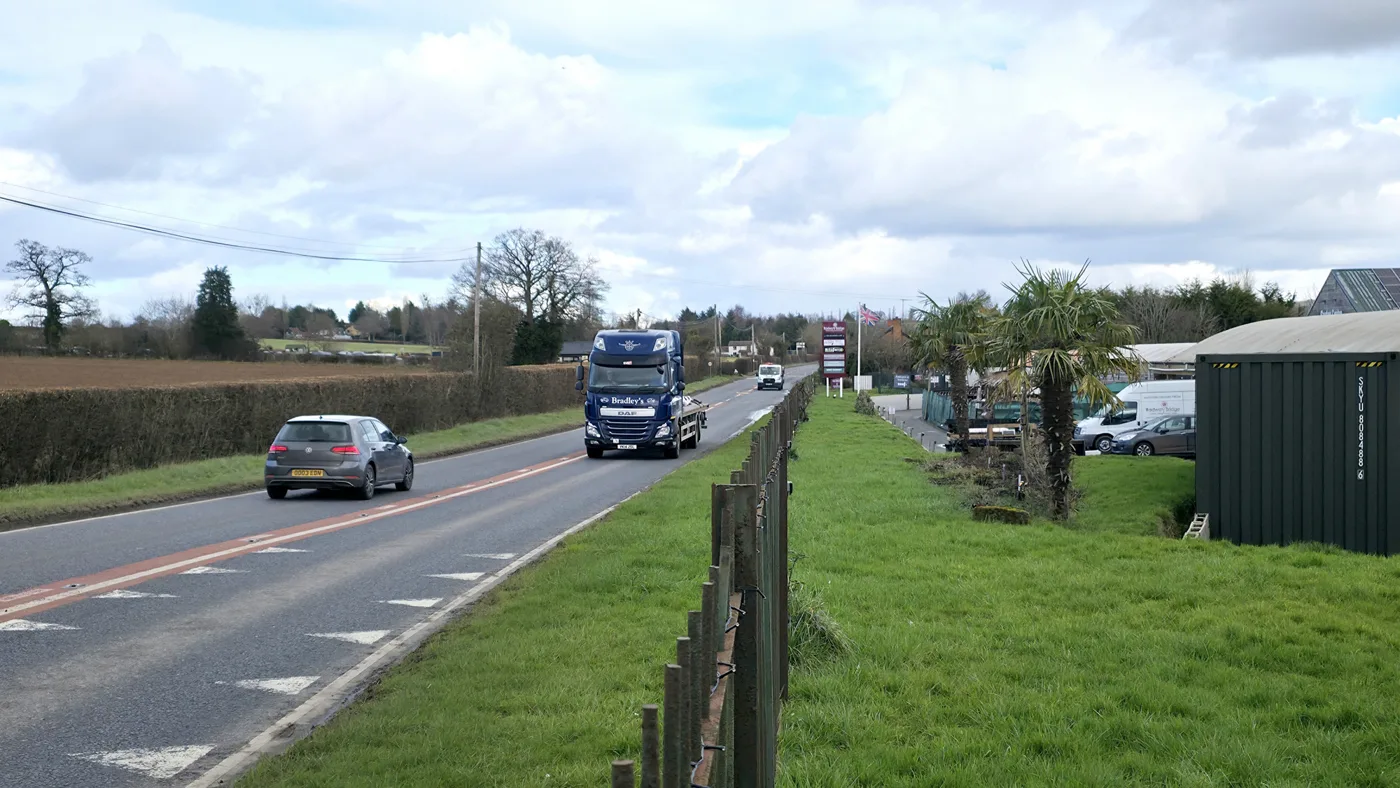 The A4103 approaching Withington from the east, with a lorry and a car on the road and a business entrance on the right